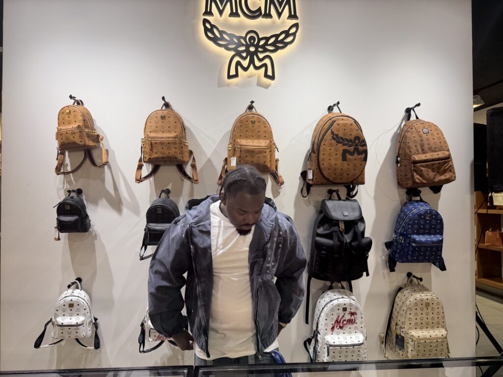 DJ A-Tron in front of the MCM logo wall of backpacks at the MCM Cabazon store, March 28, 2026
