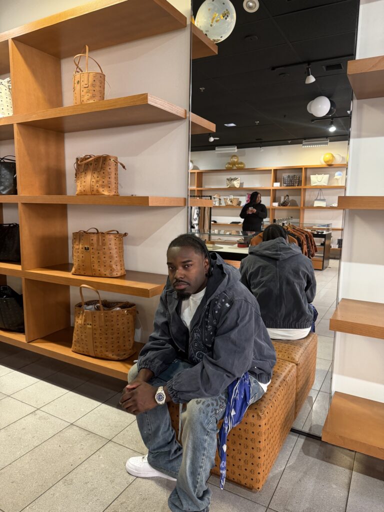 DJ A-Tron seated among MCM totes at the Cabazon store, March 28, 2026